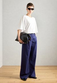 Biała, oversize koszulka, granatowe szerokie spodnie w paski, czarna skórzana kopertówka o teksturowanym wzorze oraz czarne okulary przeciwsłoneczne. Gładka drewniana podłoga.