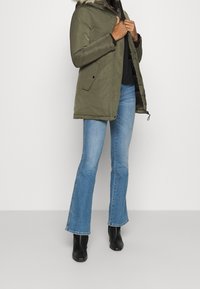 Beige parka-jacka med avtagbar pälsluva, som har framfickor, dragkedjeöppning och dragsko i nederkant, kombinerad med utsvängda jeans och boots.