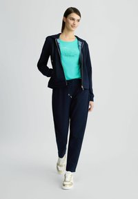 Donkerblauwe rits-hoodie met een gestructureerde afwerking, gecombineerd met een blauwe t-shirt met een bedrukt ontwerp, en donkere joggingbroek. Sneaker met gouden accenten.