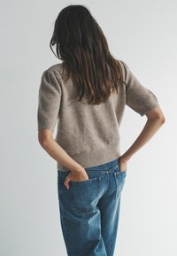 Kobieta o długich brązowych włosach, ubrana w beżowy sweter z krótkim rękawem i niebieskie dżinsy, stoi z rękami w tylnych kieszeniach na jednolitym tle.