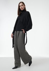 Schwarzes Strickoberteil mit übergroßen Ärmeln, kombiniert mit locker sitzenden, grauen, gemusterten Hosen. Das Model trägt schwarze spitze High Heels. Minimalistischer Hintergrund.