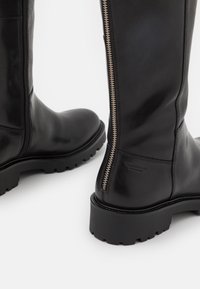 Botas de cuero negro hasta la rodilla con suela de goma gruesa, cremallera lateral y un branding mínimo cerca del talón. Textura suave y diseño aerodinámico.