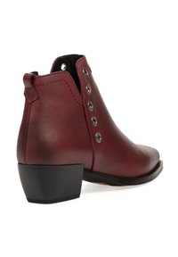 Bota de tobillo de cuero rojo con punta afilada, tacón plano negro y broches decorativos de metal plateado a lo largo del costado. Textura suave con un ligero brillo.