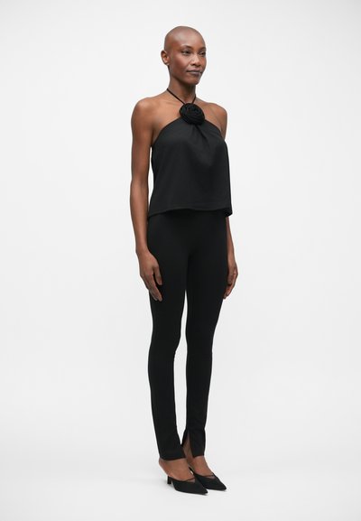 Černý crop top s květinovým akcentem na krku, spárovaný s těsně přiléhajícími černými legínami s rozparky a špičatými černými podpatky.