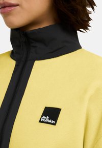 Giacca in fleece gialla con colletto a zip nero e un piccolo logo quadrato nero con testo bianco sulla parte sinistra del petto.