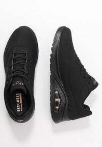 Black athletic shoes with a smooth synthetic upper, perforated details, textured sole, and visible air cushioning in the heel.