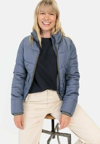 Puffetjakke i skiferblå med lynlås foran, ståkrave og ribbede manchetter, båret med en mørk top og lysfarvede bukser.