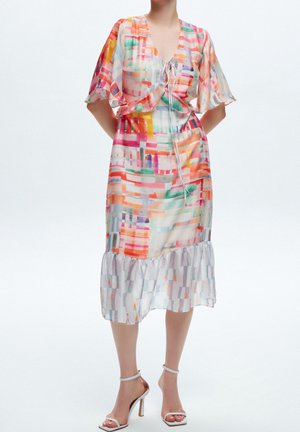 Woman wearing a colorful abstract-patterned midi dress with flutter sleeves and white high-heeled sandals on a plain background.