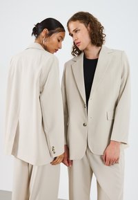 Blazers beige ajustés avec des revers en pointe et des fermetures à bouton, présentant une texture lisse. Un blazer est légèrement plus long à l'arrière.