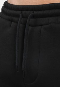 Detailní záběr na černé tepláky s elastickým pasem, šňůrkami a boční kapsou, ukazující část holé kůže nad pasem.