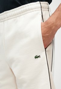 Lacoste beige sweatpants met een zwarte zijstreep, elastische tailleband en een voorzak. Voorzien van een groen geborduurd krokodillenlogo.