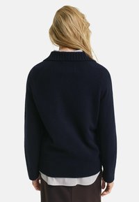 Maglione blu navy con colletto e polsini a coste, caratterizzato da un design dritto e una texture morbida. Indossato sopra una camicia a righe.