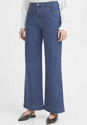 Persoon die een blauwe jeans met wijde pijpen, een witte blouse met lange mouwen en bruine platte schoenen draagt, staand tegen een effen lichte achtergrond.