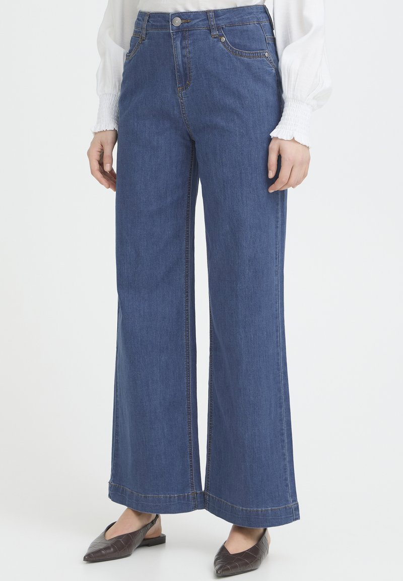 Personne portant un jean bleu à jambes larges, un chemisier blanc à manches longues et des chaussures plates marron, debout devant un fond clair et uni.