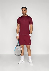 Άνδρας σε μπορντό αθλητική ενδυμασία κρατώντας ρακέτα τένις, φορώντας λευκά περιβραχιόνια, κάλτσες και αθλητικά παπούτσια, στέκεται σε απλό φωτεινό φόντο.