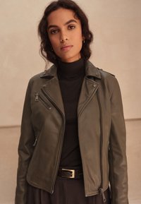 Giacca da motociclista in pelle verde oliva con cerniera asimmetrica, collo con bottone a pressione e tasche laterali, indossata sopra un dolcevita nero.