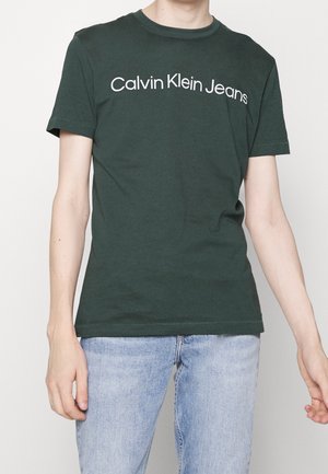 Jeune personne portant un t-shirt vert foncé Calvin Klein Jeans et un jean bleu clair, debout sur un fond clair uni.