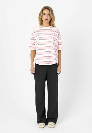 Vrouw met lang blond haar draagt een roze-wit gestreept T-shirt met korte mouwen, zwarte broek met wijde pijpen en witte sneakers.