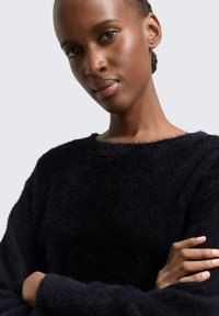 Schwarzer, flauschiger Pullover mit Rundhalsausschnitt, strukturiertem Stoff und langen Ärmeln. Das Modell hat die Arme verschränkt und zeigt so die Passform und Textur des Kleidungsstücks.