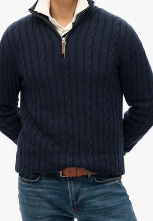 Homme portant un pull en maille torsadée bleu marine à fermeture éclair quart de zip par-dessus une chemise blanche à col, associé à un jean bleu et une ceinture en cuir marron.