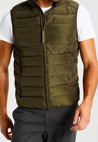 Olivgrüße, mit Reißverschluss versehene Weste mit einem gepolsterten Design und horizontaler Absteppung. Über einem weißen T-Shirt getragen, aus leichtem Material.