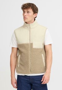 Beige und braune ärmellose Weste aus Fleece und glattem Stoff, mit hohem Kragen, Fronttasche und seitlichen Reißverschlusstaschen.