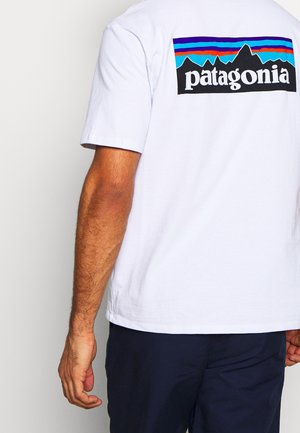 Mann trägt ein weißes Patagonia-T-Shirt mit mehrfarbigem Berglogo auf der Rückseite, kombiniert mit dunklen Shorts vor schlichtem Hintergrund.