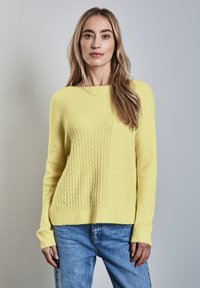 Gul stickad tröja med ett strukturerat mönster med varierande ribbor. Matcher med blå denimjeans, designen är lös och bekväm.