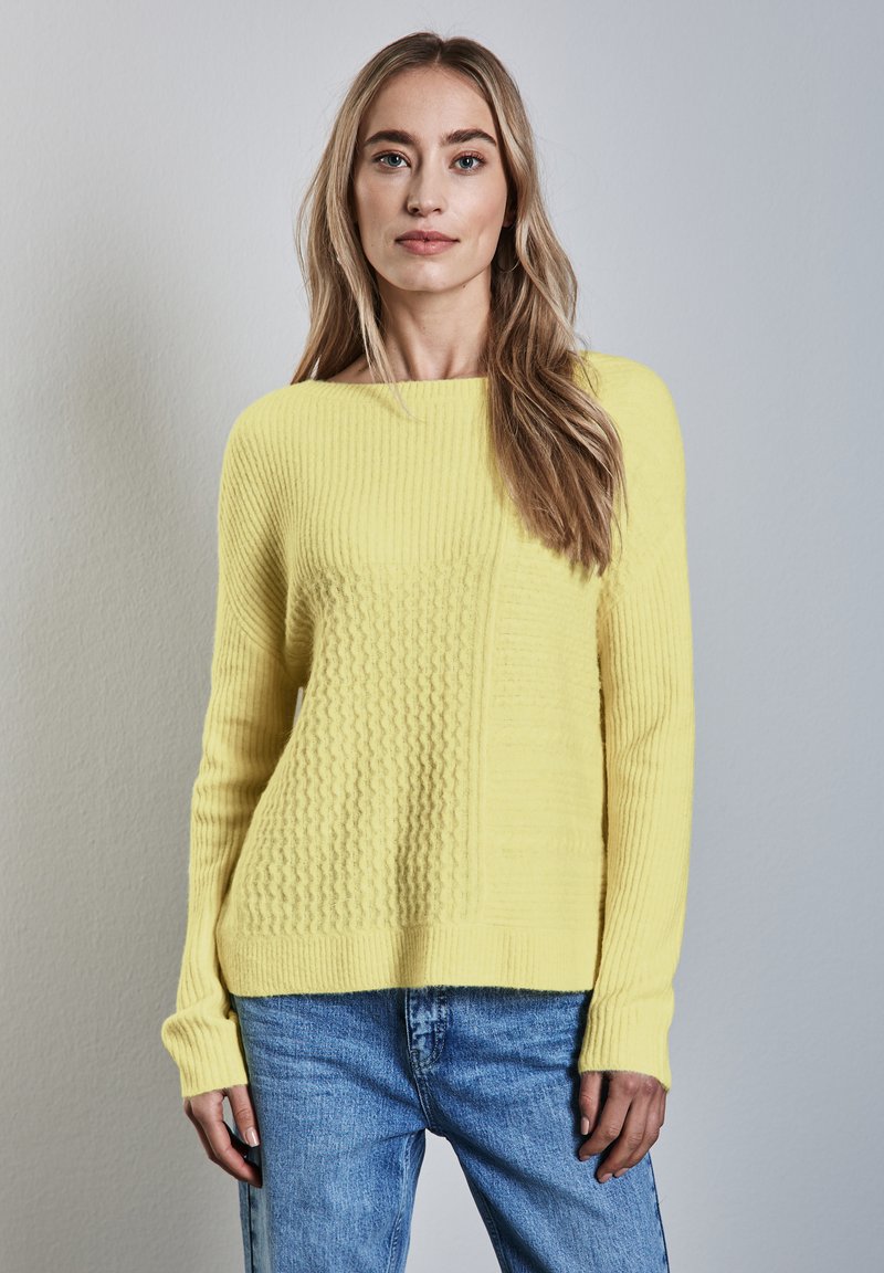 Gul stickad tröja med ett strukturerat mönster med varierande ribbor. Matcher med blå denimjeans, designen är lös och bekväm.