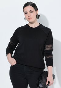 Felpa nera con maniche lunghe che presenta dettagli in mesh trasparente e scollatura a girocollo. Indossata con pantaloni neri e in mano tiene una pochette nera.