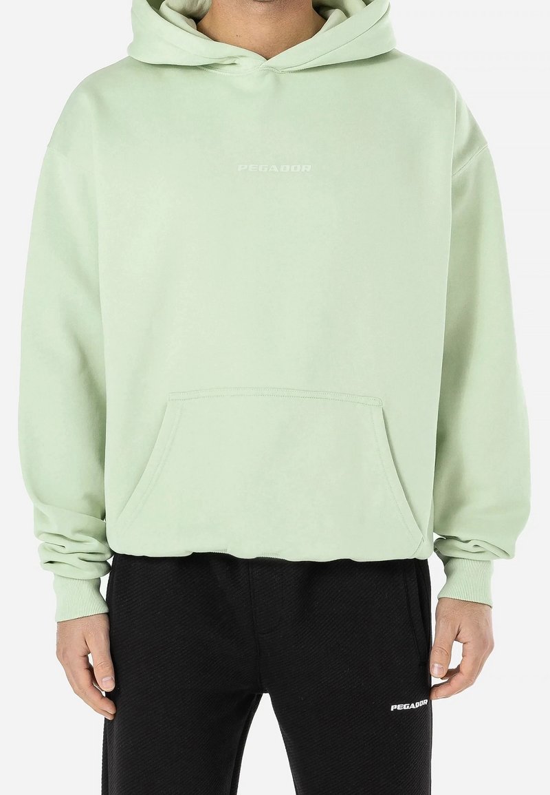 Homme portant un sweat à capuche vert clair avec poche avant et un pantalon noir, tous deux avec le logo "PEGADOR", debout les bras détendus le long du corps.