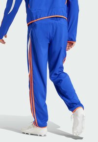 Blå träningsbyxor med orange accenter, har sidostriper, en avslappnad passform och avsmalnande ben. Materialet verkar mjukt och lättviktigt.