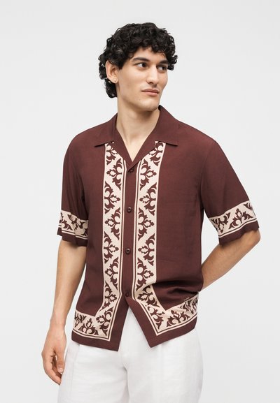 Νεαρός άνδρας που φοράει καφέ κοντομάνικο πουκάμισο με μπεζ λουλουδένια σχέδια και λευκό παντελόνι, κοιτάζει προς τα αριστερά του.