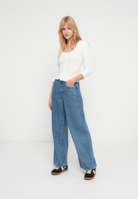 Νεαρή γυναίκα που στέκεται με το ένα χέρι στην τσέπη, φορώντας λευκό μακρυμάνικο πουκάμισο με κουμπιά, παντελόνι καμπάνα μπλε και μαύρα αθλητικά παπούτσια.