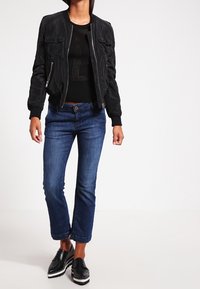 Schwarze Bomberjacke mit Reißverschlusstaschen, kombiniert mit einem schwarzen Stricktop, blauen, kurzen Jeans und schwarzen Plateau-Schuhen mit weißen Sohlen.