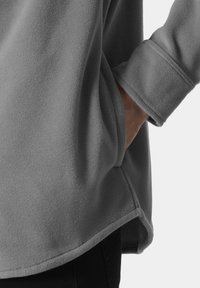 Szary polarowy sweter o luźnym kroju, z zaokrąglonym dołem i widocznymi szwami na rękawach. Miękki, fakturowany materiał.