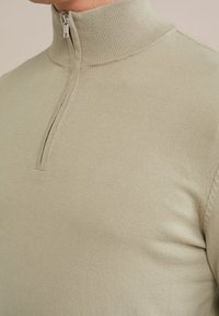 Närbild av en person som bär en beige stickad tröja med en ribbad ståkrage och en kort silverfärgade dragkedja vid halsen.