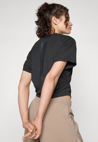 T-shirt nera a maniche corte realizzata in tessuto morbido, con una striscia lucida verticale sulla parte posteriore. Abbinata a pantaloni beige con vita elastica.