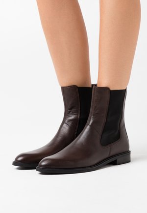 Hnědé kožené kotníkové boty s elastickými bočními panely nošené na holých nohách, zobrazené na bílém pozadí.