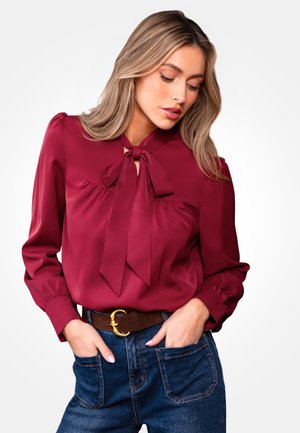 Burgundowa satynowa bluzka z kokardą przy dekolcie, bufiastymi rękawami i luźnym krojem, skomponowana z wysokim stanem dżinsów i brązowym paskiem.