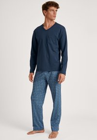 Marineblauwe langekleurige V-hals top met zak, gecombineerd met blauwe pyjamabroek met een geometrisch patroon. Model staat blootsvoets.
