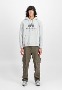 Lichtgrijze hoodie met een voorzak, voorzien van een zwart logo; gecombineerd met olijfgroene cargobroek en grijze sneakers met een patroon.