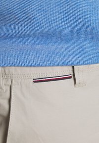 Pantalones beige con una textura suave, que presentan un acento de rayas rojas, blancas y azules cerca de la cinturilla y una camisa de patrón azul claro arriba.