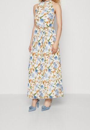 Kobieta ubrana w bezrękawnikową, kwiecistą suknię maxi w niebieskie, pomarańczowe i zielone kwiaty, zestawioną z niebieskimi butami na wysokim obcasie z kwiatowym detalem.