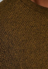 Strickpullover in Dunkelgoldbraun mit einem strukturierten, gerippten Muster. Eine Nahaufnahme zeigt die filigrane Naht und das weiche Erscheinungsbild des Stoffes.