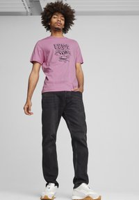 Helles rosa T-Shirt mit Grafikdruck "PUMA BOWL", kombiniert mit dunklen Jeans und weißen Sneakers mit mehrfarbigen Akzenten.