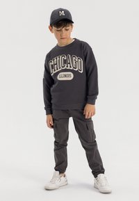 Donkergrijze sweatshirt met "Chicago Illinois" in beige gedrukt. Gecombineerd met donkergrijze cargobroek en witte sneakers. Pet met de letter "M."