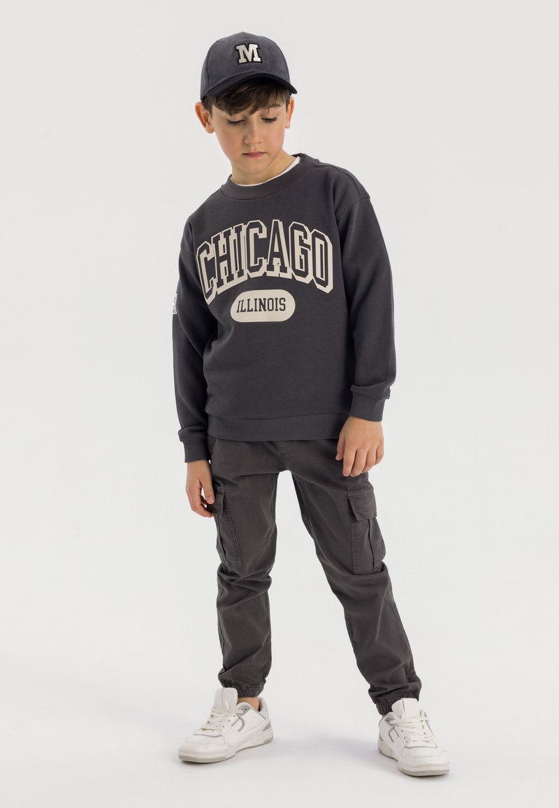 Donkergrijze sweatshirt met "Chicago Illinois" in beige gedrukt. Gecombineerd met donkergrijze cargobroek en witte sneakers. Pet met de letter "M."