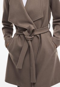 Abrigo marrón de tela suave con cinturón en la cintura, que presenta profundos bolsillos laterales y un diseño de solapa amplia. Diseñado para un ajuste cómodo.