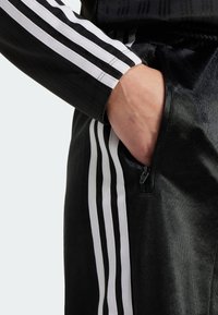 Black athletic pants with white stripes, featuring a zippered pocket and a smooth, shiny texture. The design includes vertical lines and a fitted silhouette.